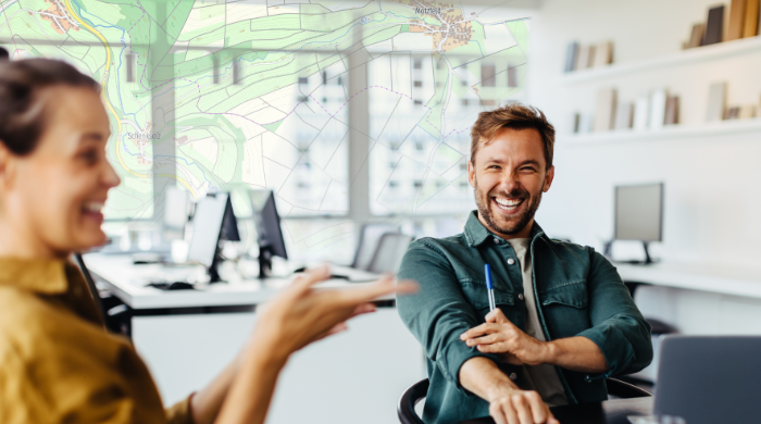 Kreative Teamarbeit und Geodaten – Kartenbasierte Lösungen im modernen Büro Zwei lachende Kollegen in einem modernen Büro mit Computern, im Hintergrund eine topografische Landkarte